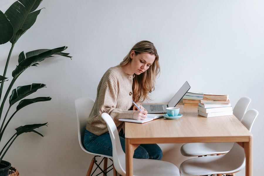 Woman writing in a notebook at a desk with a laptop, batching work at home