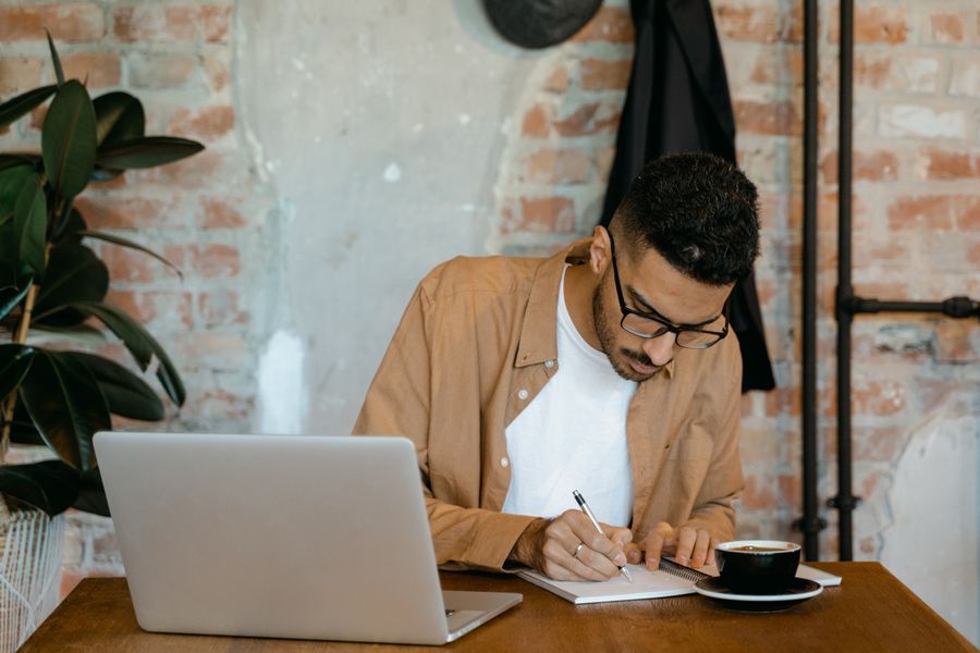 Man writing in a notebook at a desk with a laptop and a cup of coffee