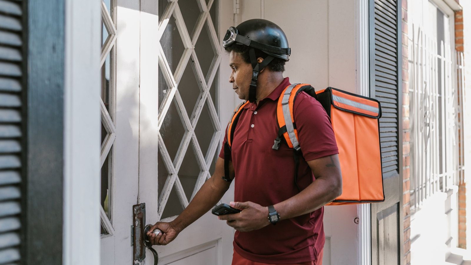 Food delivery worker in a helmet with an orange insulated delivery backpack, holding a phone at a customer's front door