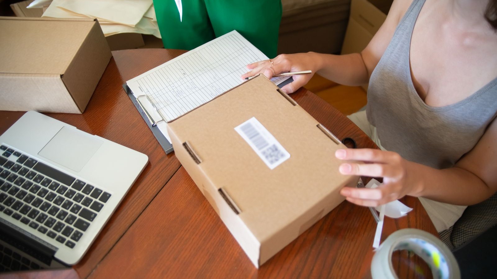 Woman sealing a labeled cardboard shipping box at a wooden desk, with a laptop and an order clipboard beside her