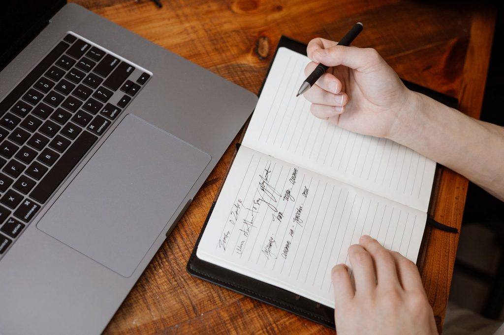 A hand writing notes in a notebook next to a laptop while verifying a job listing